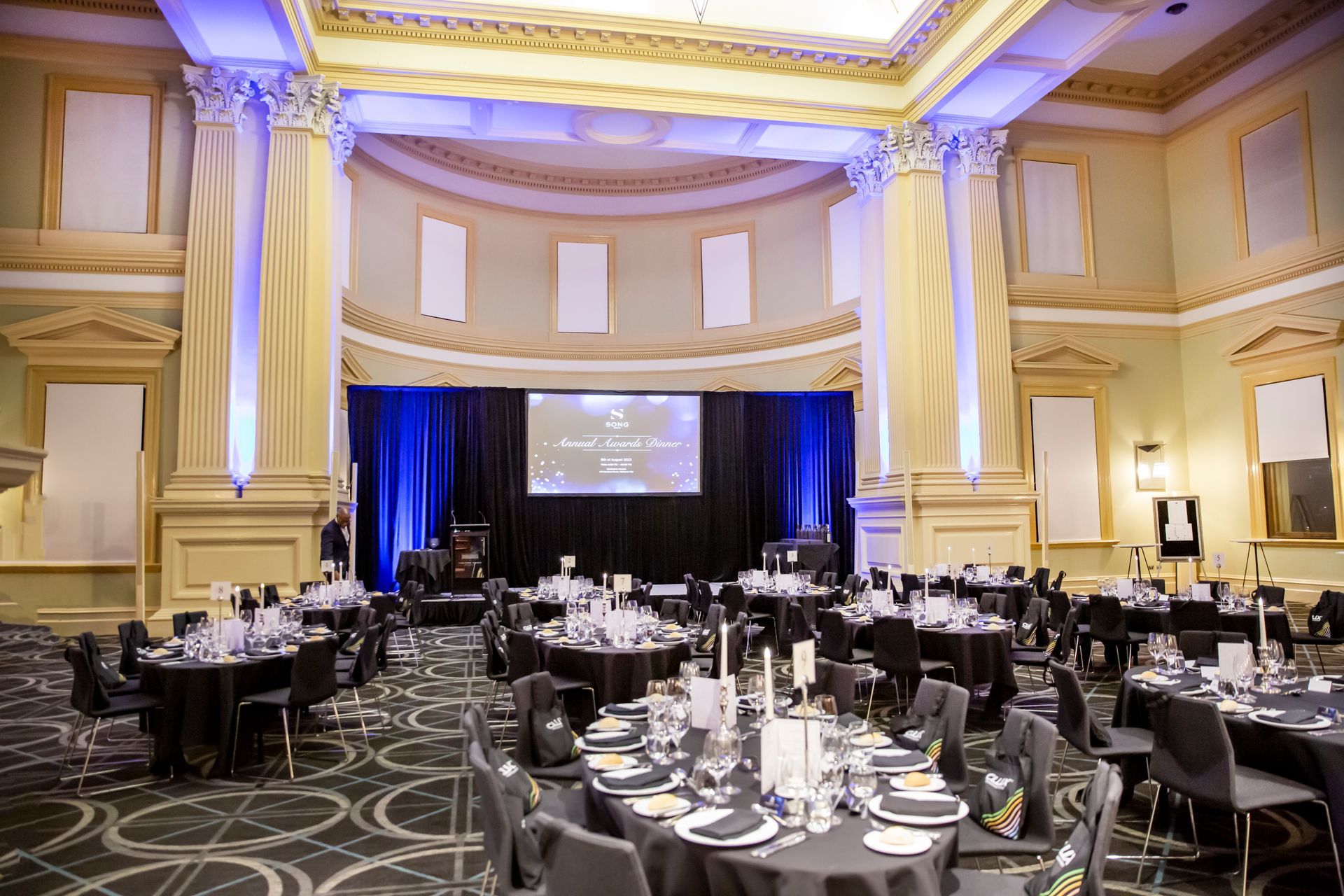 Elegant ballroom set for an event with round tables, black linens, and a stage with a screen.