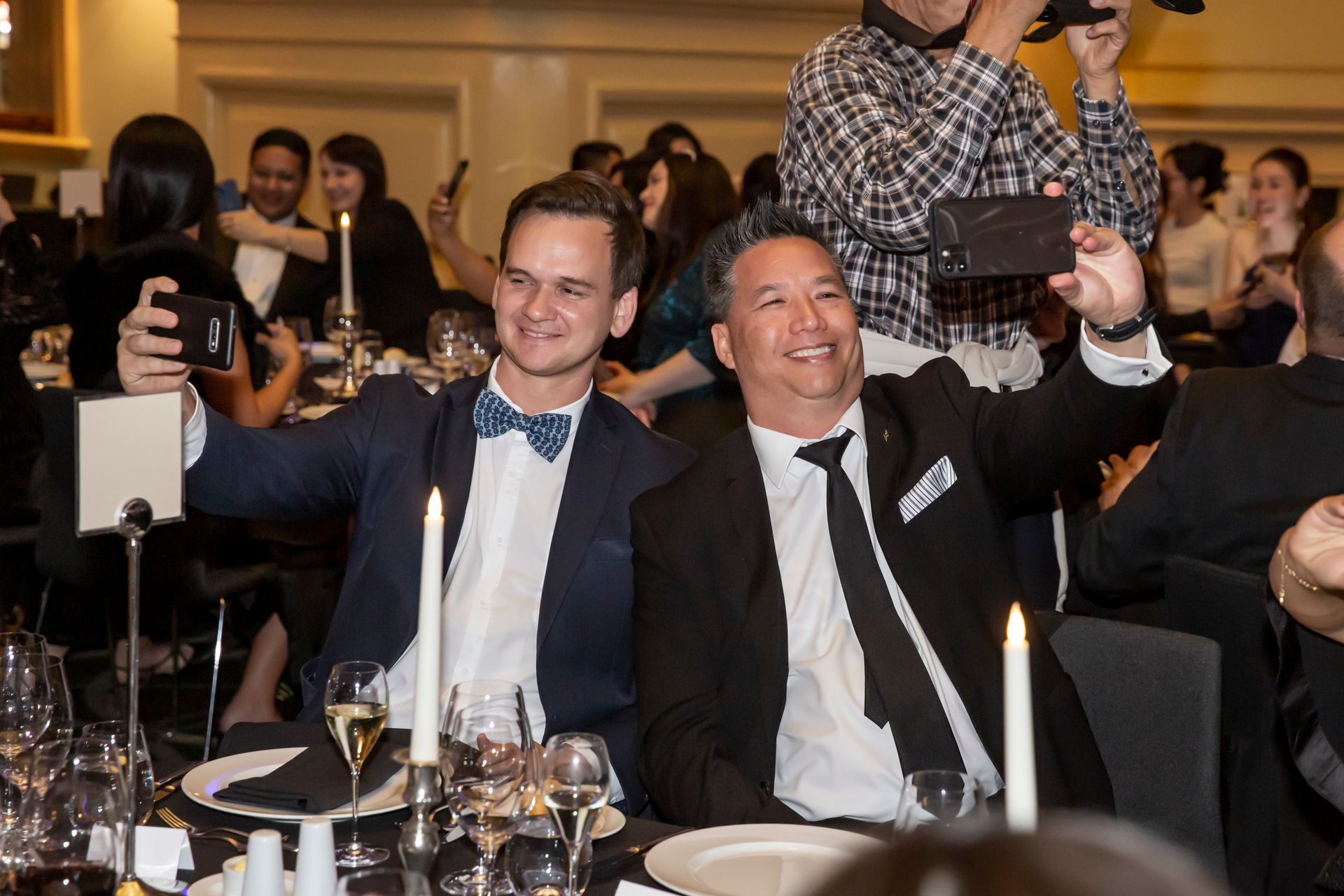 Two men at a formal event taking a selfie with smartphones. They are smiling. Formal attire, candlelight, and a crowd.