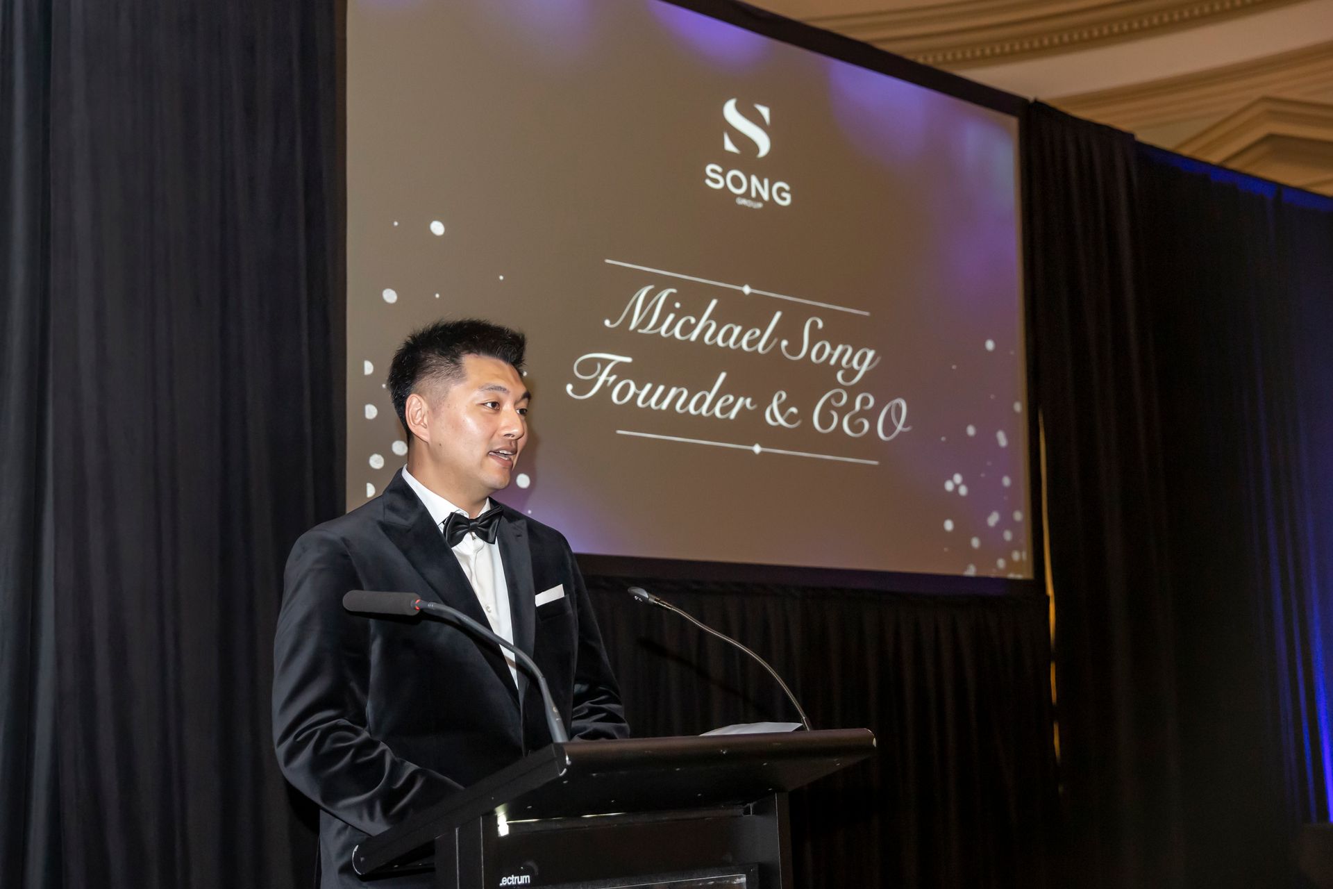 Michael Song, Founder & CEO, speaking at event; wearing a tuxedo. Black background with logo.