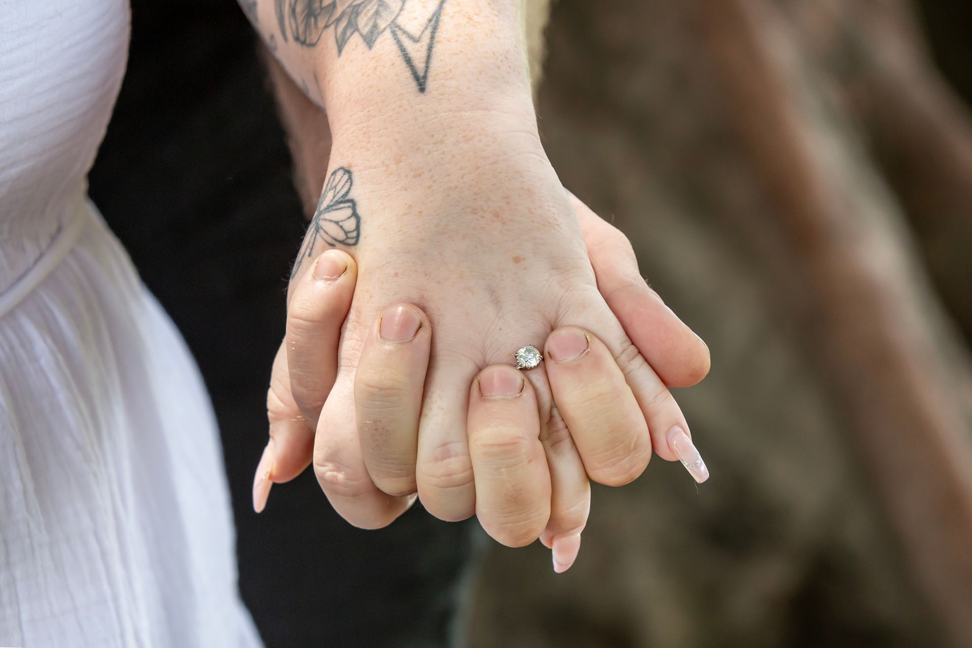 Couple holding hands, one with an engagement ring. Tattoos on the woman's hand, wearing a white dress.