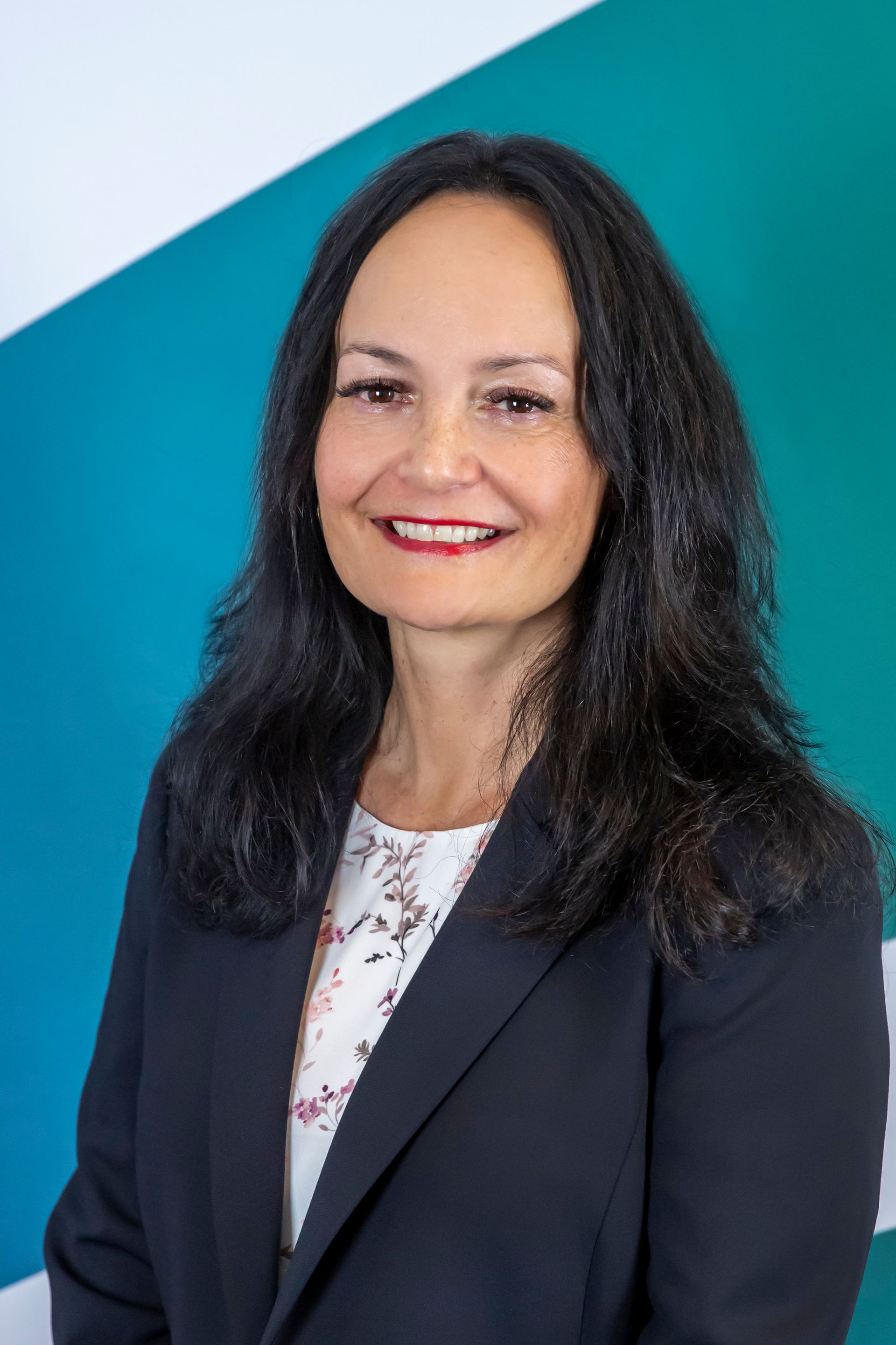 Woman with long dark hair, wearing a black blazer and patterned blouse, smiling. Teal and white background.