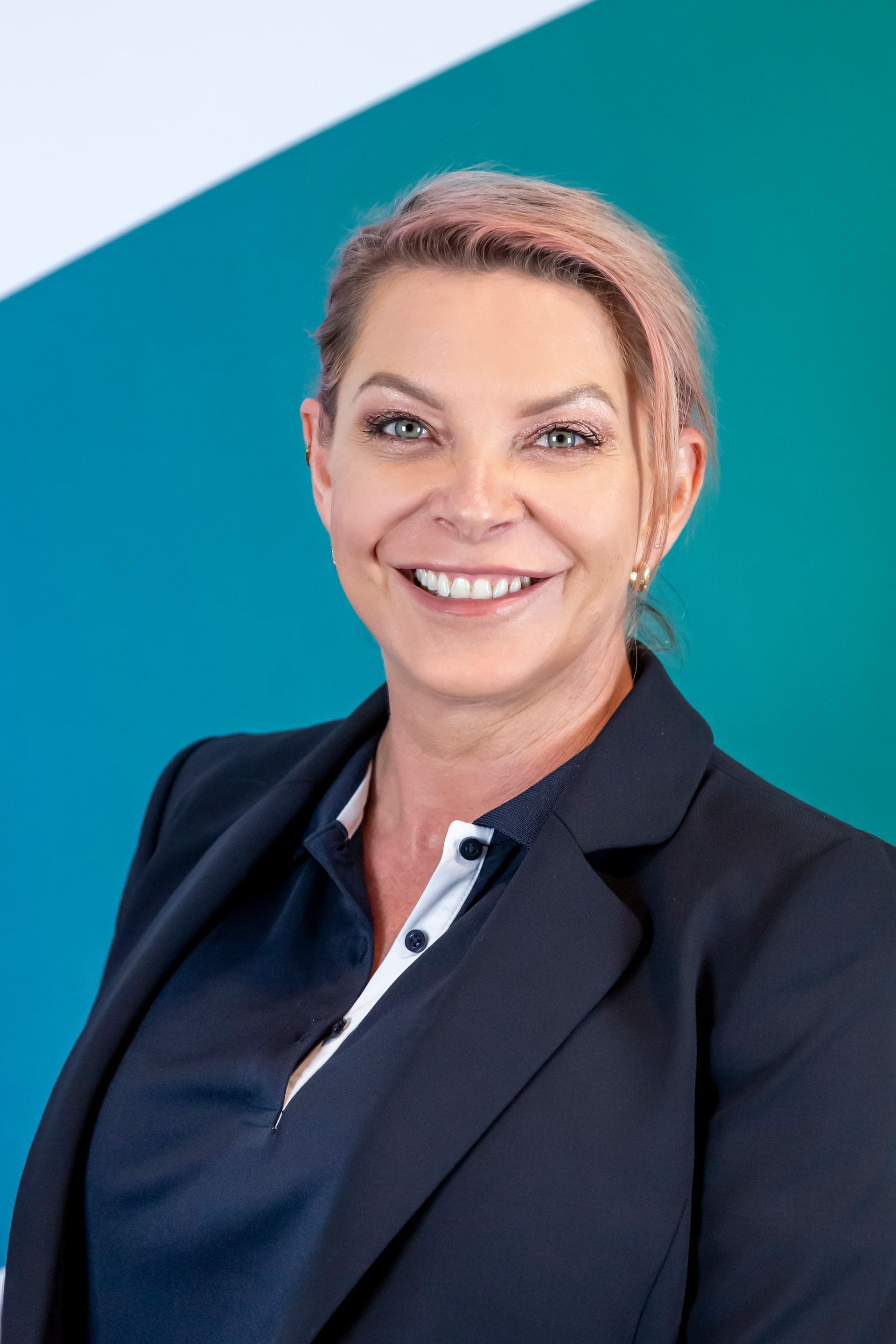 Woman with pink-tinged hair smiles at the camera, wearing a blazer and polo shirt against a teal backdrop.