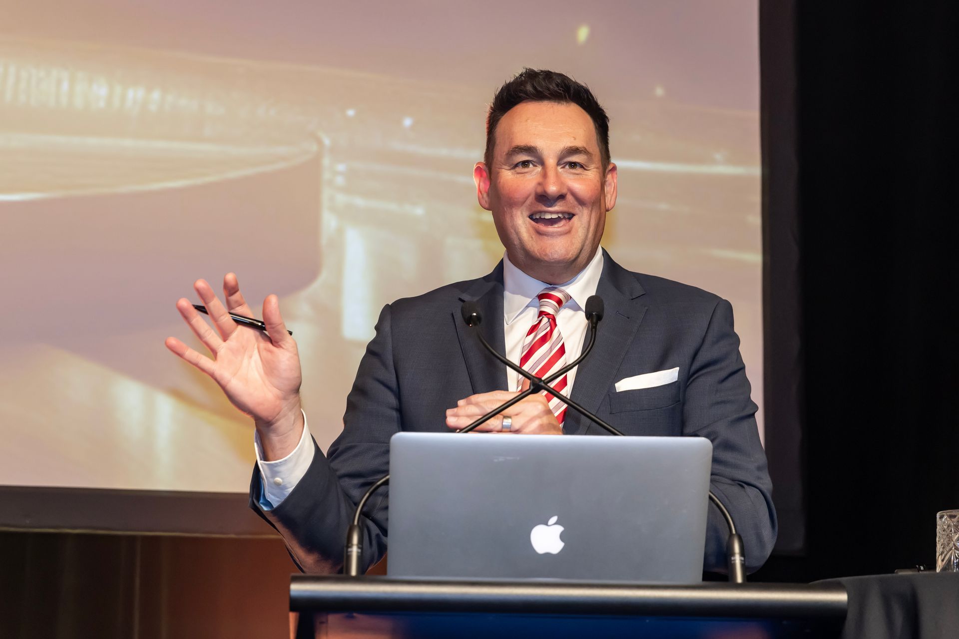 Man in suit speaking at a podium, gesturing with a hand; laptop visible.
