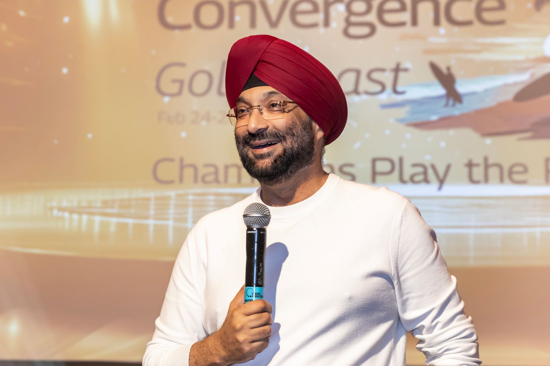 Man in red turban speaking at a conference, holding a microphone, smiling.