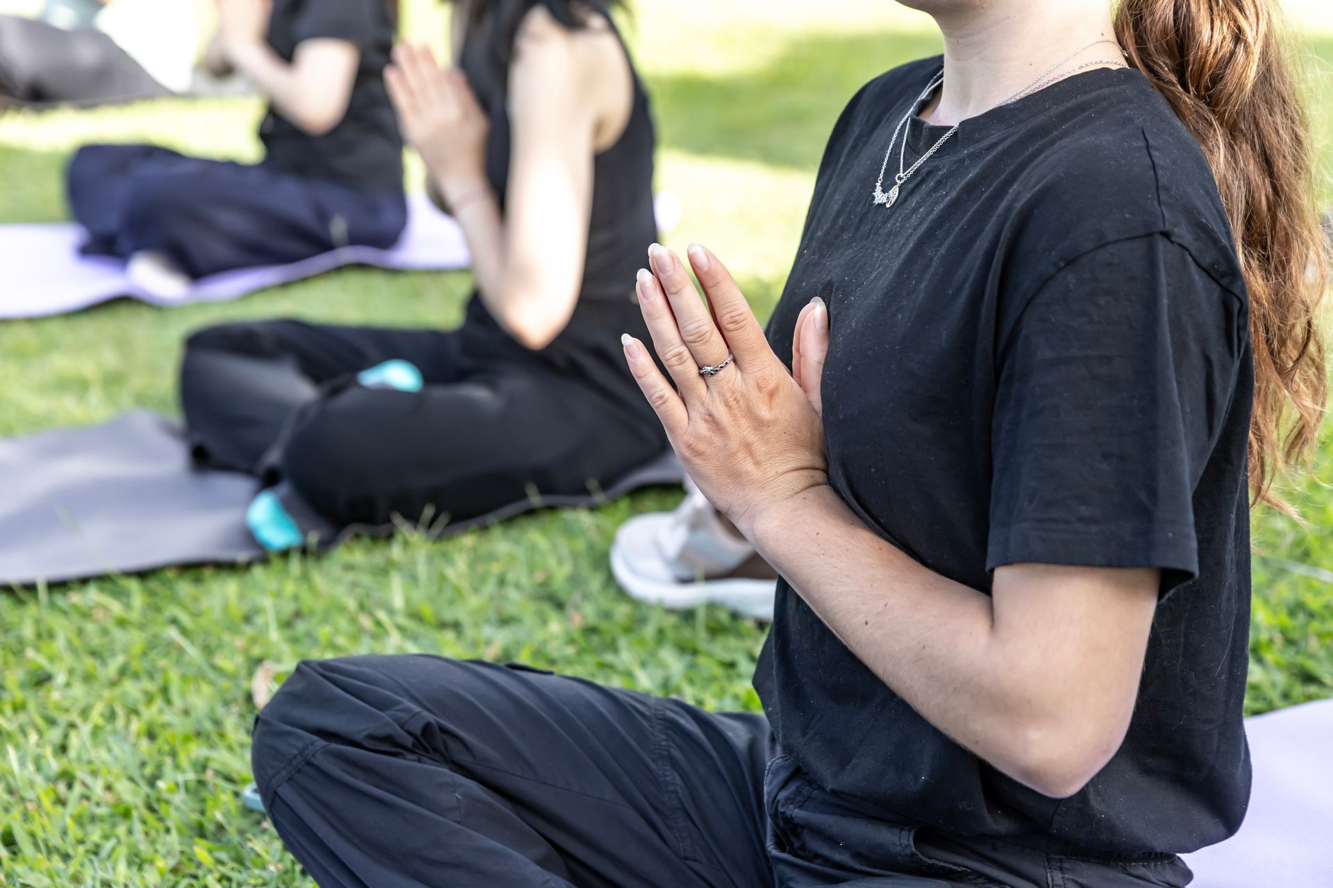 People in a park doing yoga, hands in prayer pose, wearing black clothing, sitting on mats.