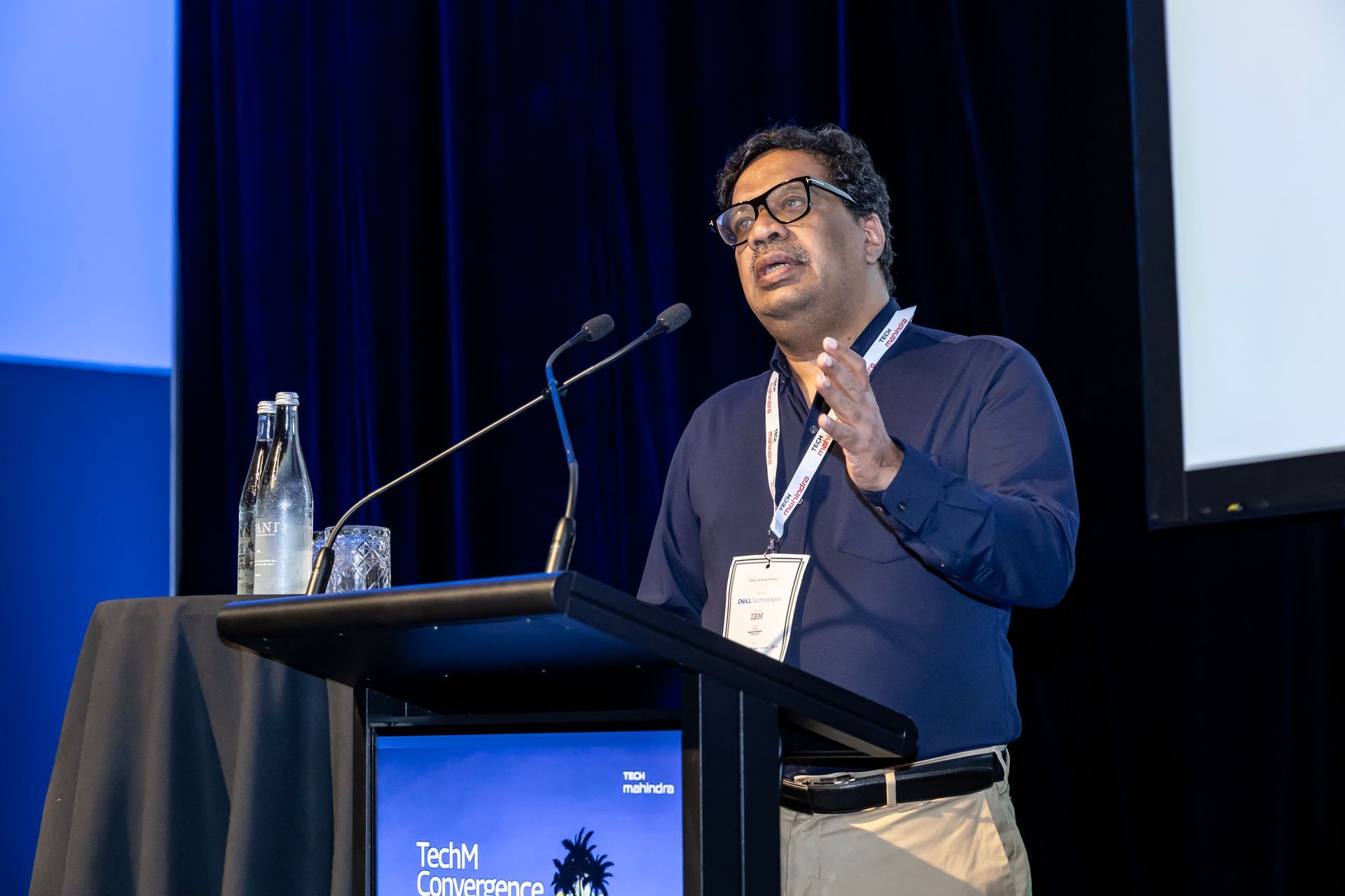 Man speaking at a podium, gesturing with his hand. He wears glasses, a blue shirt, and tan pants. Conference setting.
