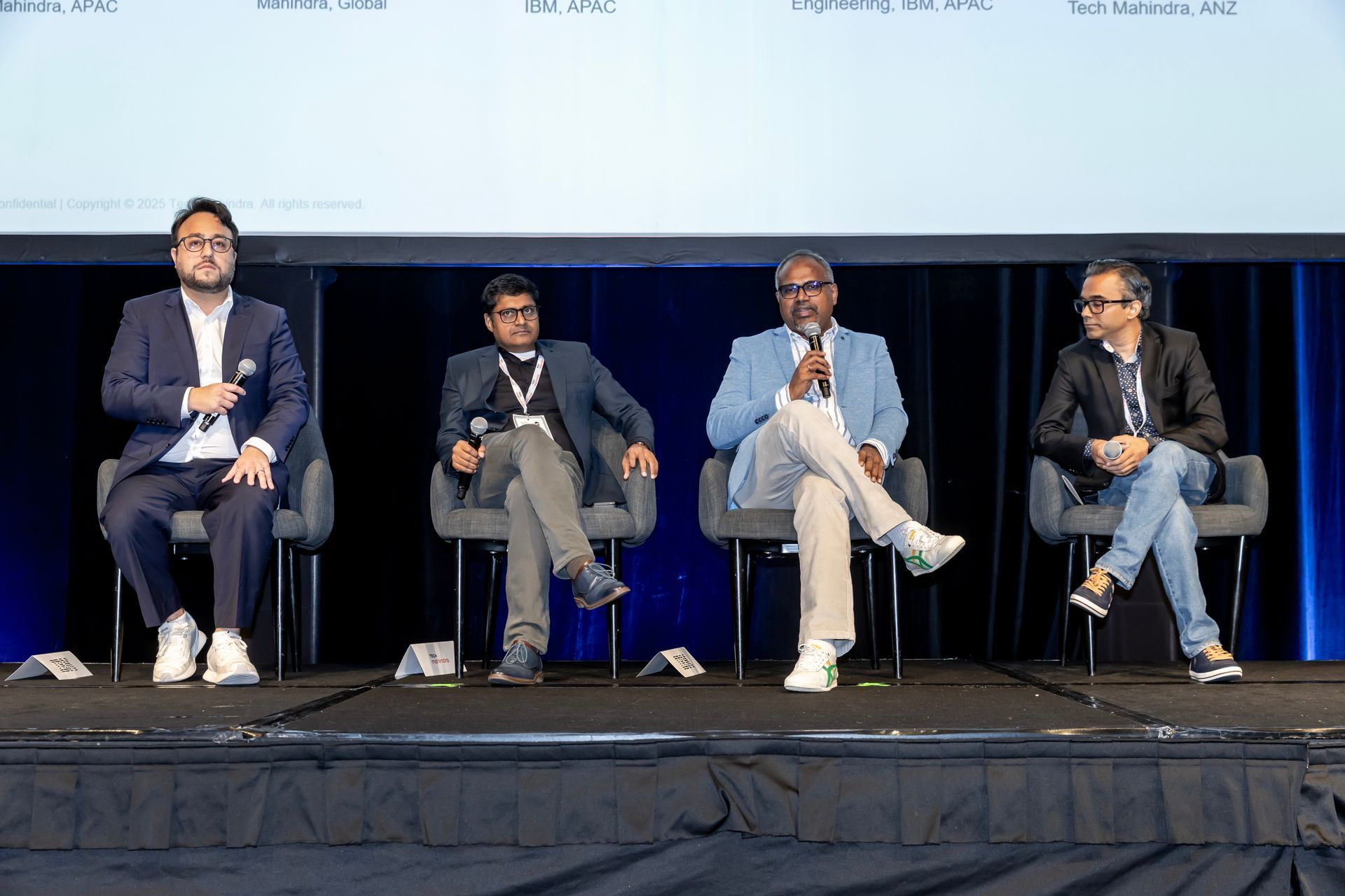 Four men in suits sit on stage at a conference, speaking into microphones.