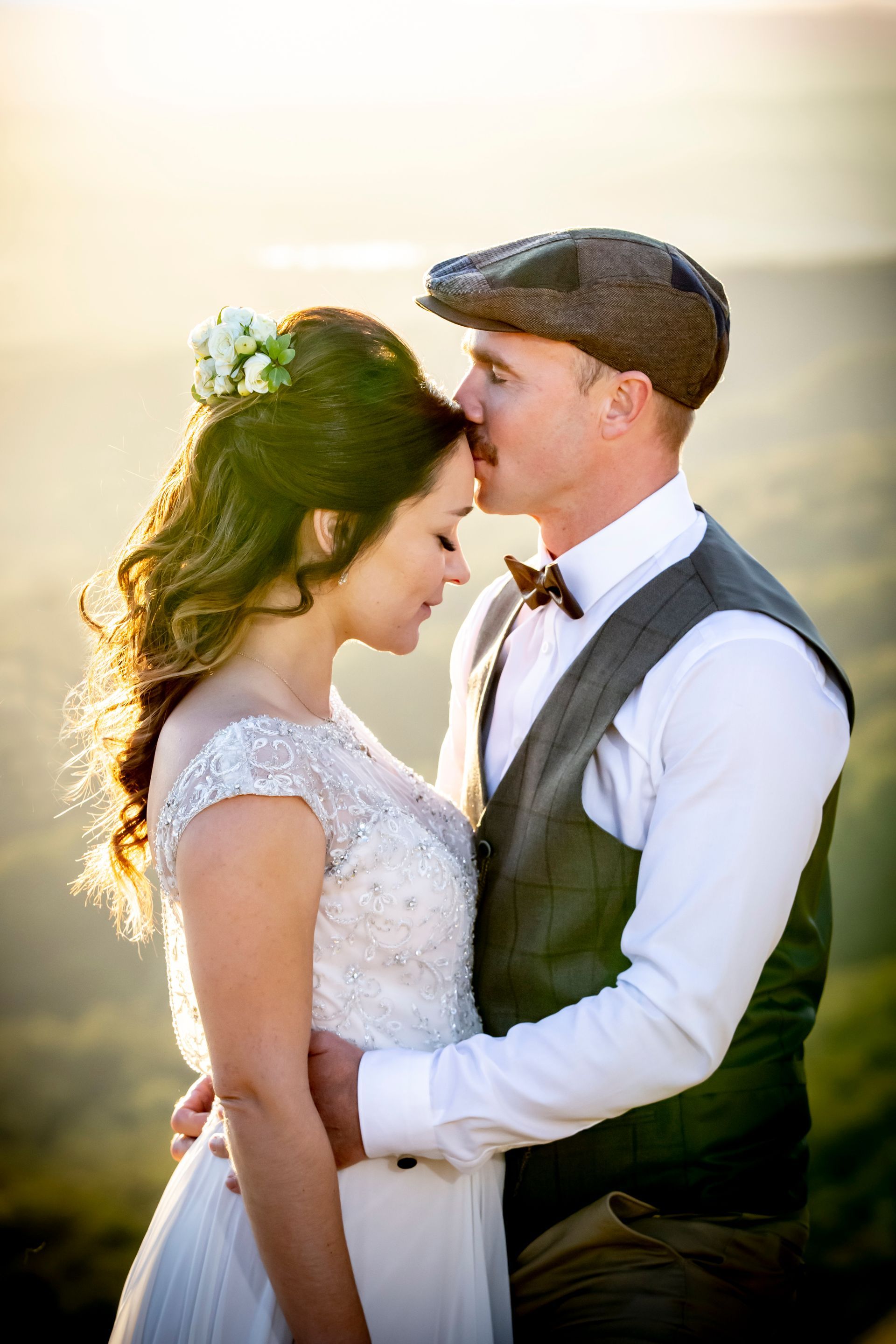 Bride and groom in embrace, man kissing woman's forehead at sunset, wearing vintage attire. Photography by Andy Williams