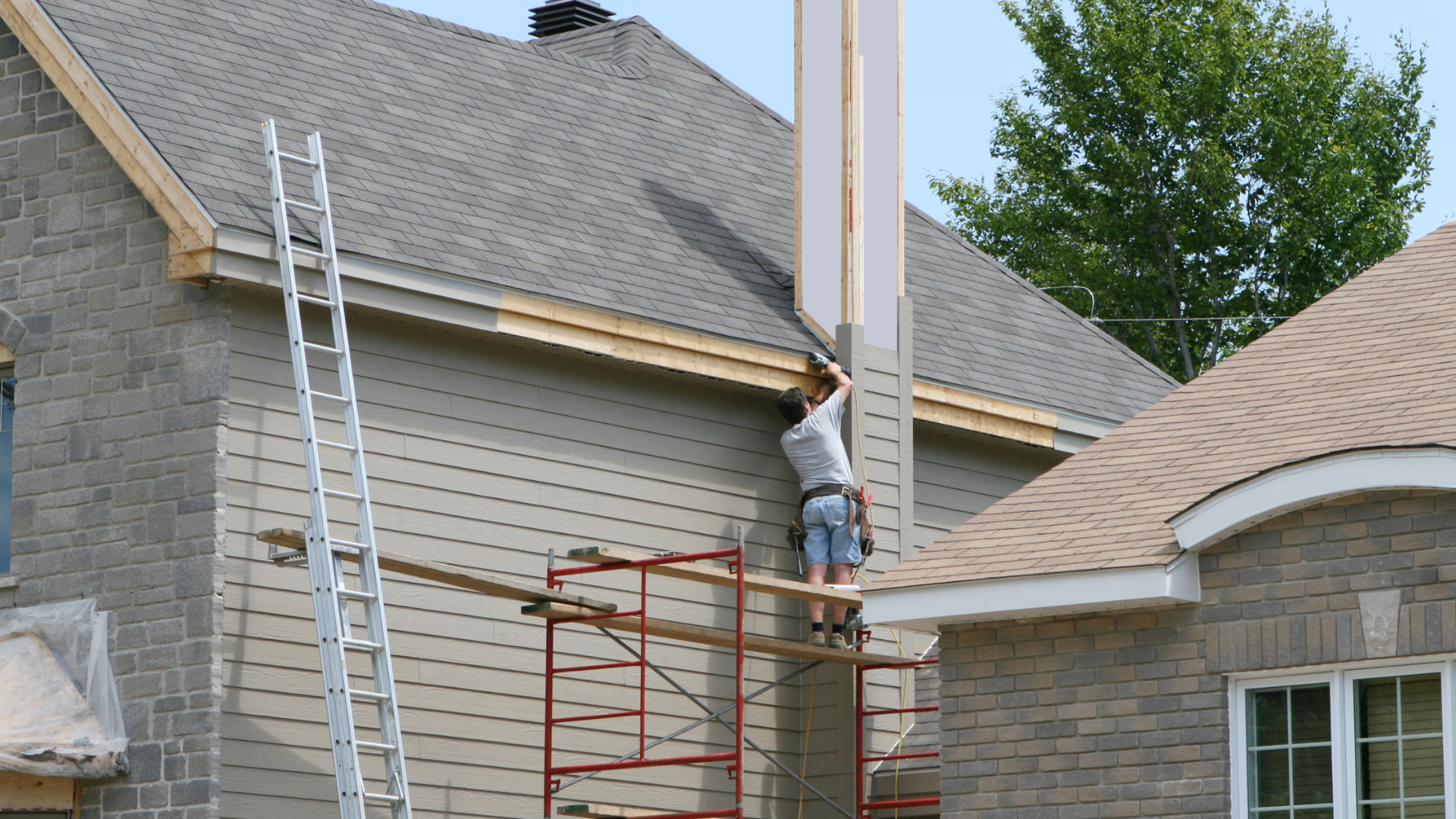 A construction worker installing siding on a house, standing on scaffolding near a chimney.