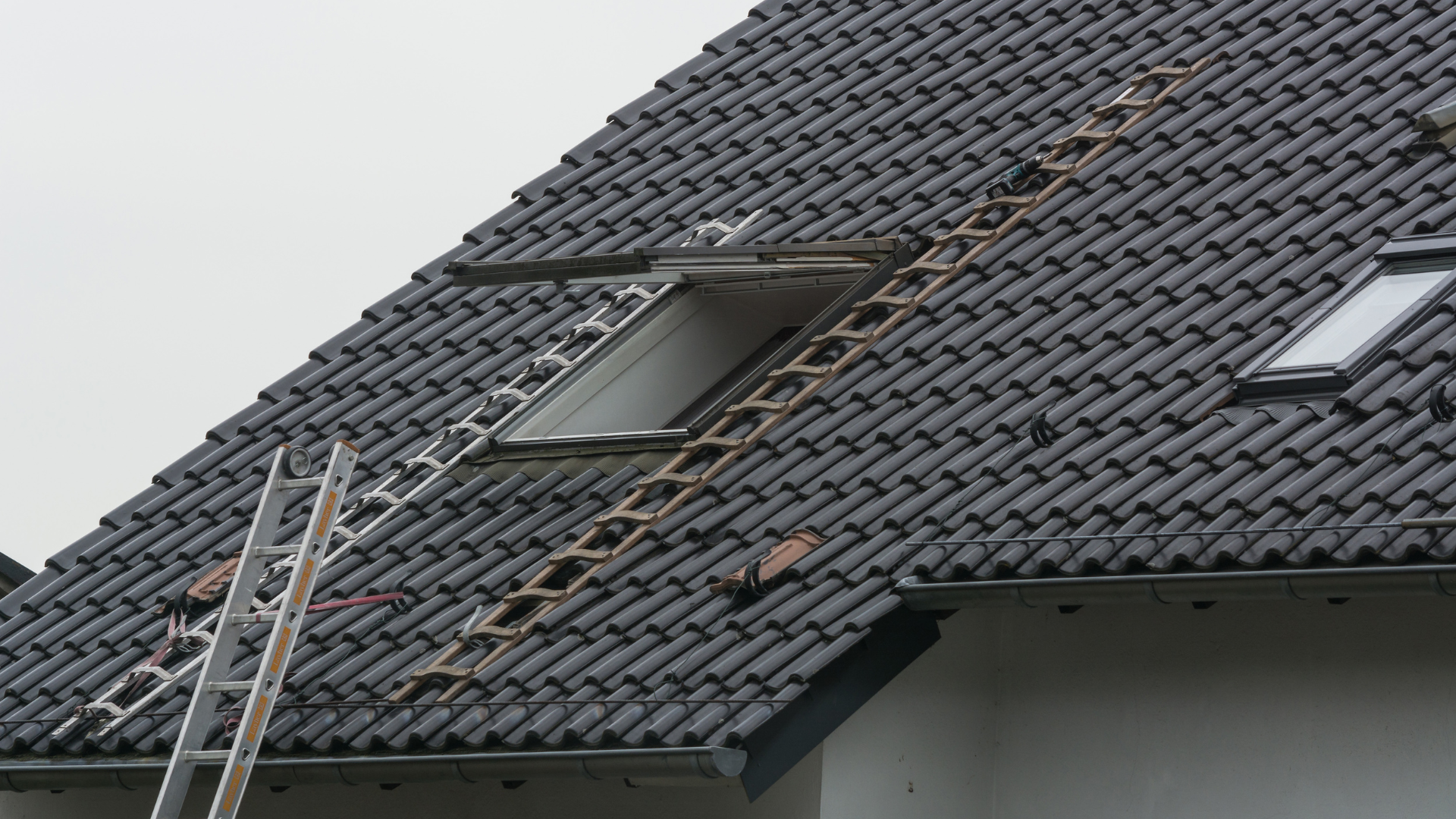 Two ladders on a dark tiled roof; one leads to an open skylight, one to the ground.
