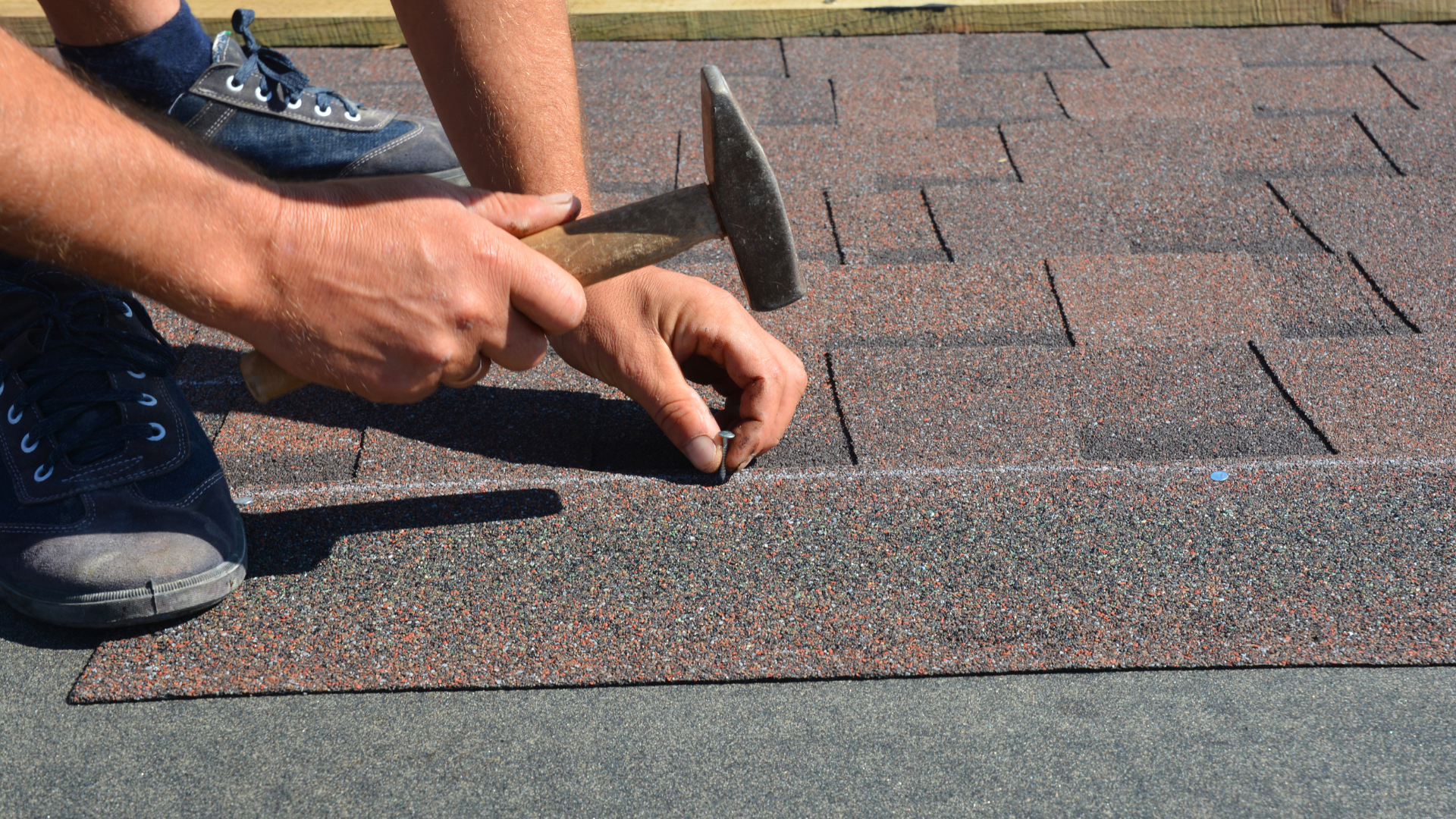 Person hammering a nail into a roofing shingle, outdoors.