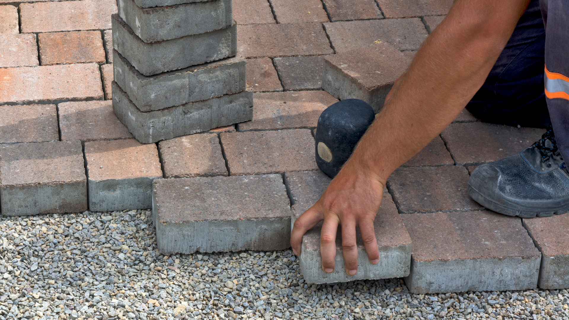 Person placing brick pavers, constructing a walkway; grey and reddish-brown bricks with gravel base.