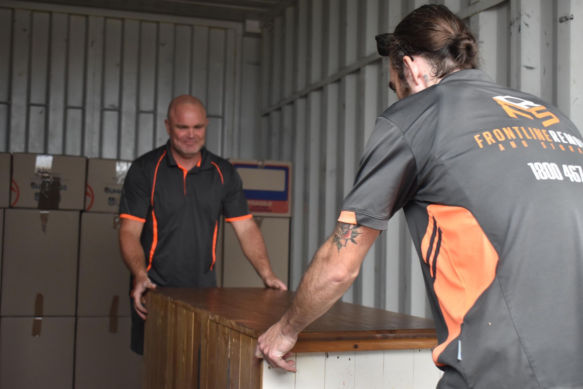Two men loading a wooden cabinet into a storage unit. They wear gray and orange shirts — Frontline Removals And Storage in Port Macquarie, NSW
