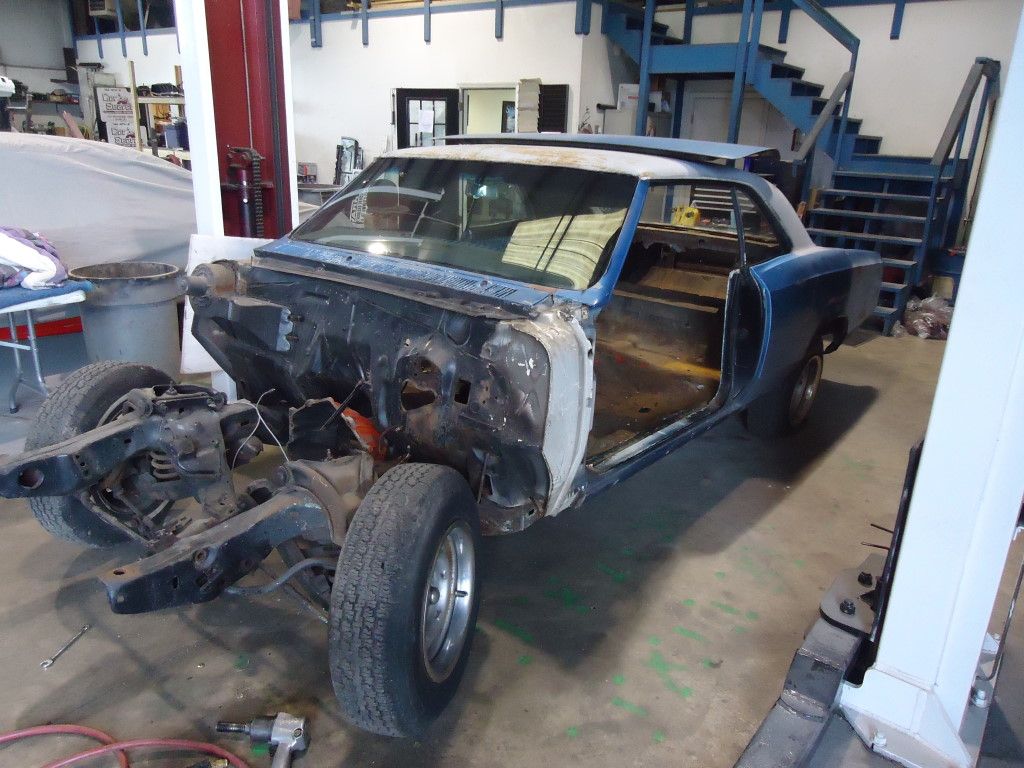 A partially disassembled blue classic car inside a workshop, with the front frame and wheels detached.