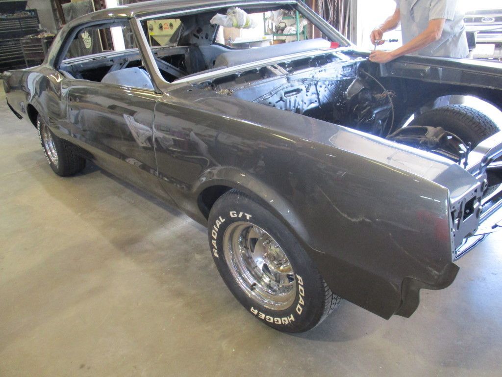 Dark gray classic car undergoing restoration in a workshop; a person works on the hood.