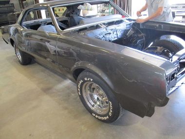 Dark gray classic car undergoing restoration in a workshop; a person works on the hood.