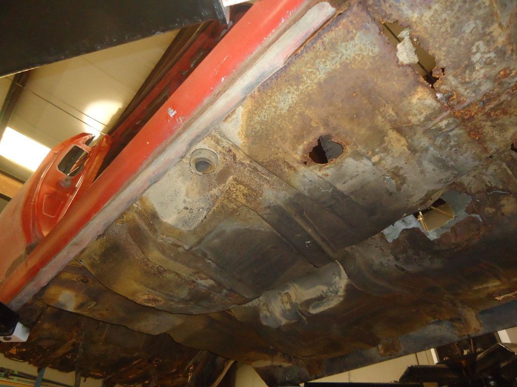 Underside of a rusty red car, showing extensive corrosion and holes in the floorboards and undercarriage.