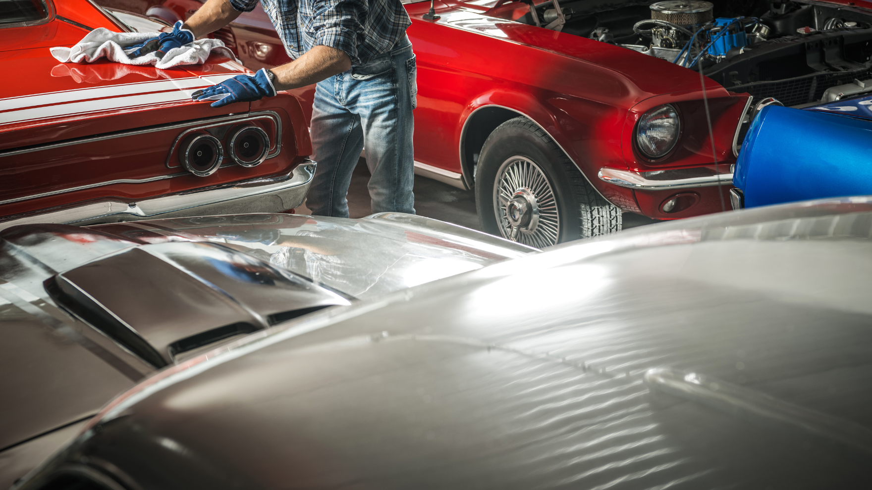 Person cleaning classic cars, including a red one with white stripes. Setting is a garage with other vehicles.