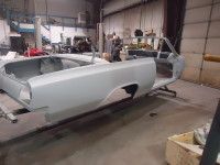 Car body in a repair shop; gray, disassembled, with a person working in the background.