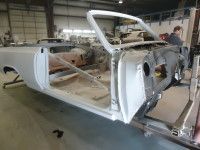 A light gray, classic car body in a workshop, under restoration. A person is working on the car.