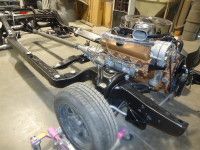 Black car chassis with engine, transmission, and front wheel in workshop.