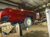 A maroon classic car in a shop, elevated on a lift. The wheels are yellow.