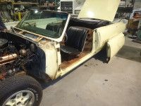 Yellow convertible car undergoing restoration in a garage, missing parts, hood and door open.