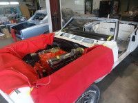White convertible car in a workshop, engine exposed, covered in red protective wrapping. Another car in background.