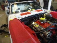 White classic car, engine exposed, covered in red, under repair in a garage.