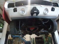 Underside of a classic car on a lift; silver bumper, black tires, and work setting visible.