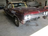 Burgundy classic car missing front bumper and grill, in a garage.