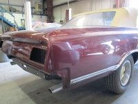 Burgundy convertible car in a workshop, gold top, rear view, exhaust pipes visible.