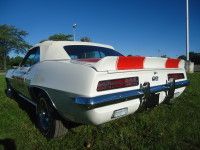 White 1969 Chevrolet Camaro convertible with red and white stripe spoiler, parked on grass against a blue sky.