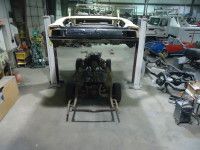 A car chassis raised on a lift, engine on a dolly in a garage.