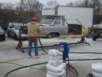 Man sandblasting a vintage car's body in an outdoor lot, wearing safety gear; blue sandblasting machine present.
