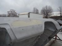 Gray car body under repair outdoors with a cloudy sky and bare trees in the background.