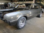 Classic gray car in a body shop, mid-restoration with no engine components and white-lettered tires.
