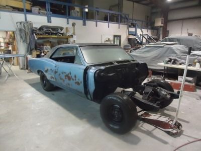 Classic blue car in a workshop, mid-restoration with missing front end, black interior, and dark wheels.