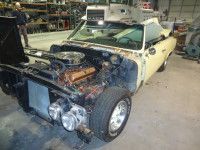 Yellow classic convertible car under repair in a garage, missing front body panels, hood open, engine visible.