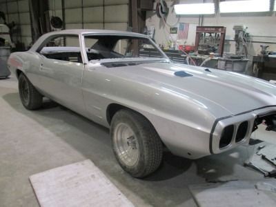 Silver 1968 Pontiac Firebird coupe in a workshop, undergoing restoration. The car lacks glass and trim, showing primer.
