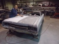 Man standing near a convertible car in a workshop; car is partially painted in two shades of gray.