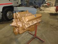 Gold-painted car engine on a red stand in a workshop. A truck and vehicle parts are in the background.
