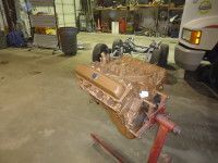 Brown engine on a stand in a garage. A car frame is in the background.