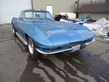 A classic blue Corvette sports car parked on wet asphalt. It's a sunny day with some snow visible in the background.
