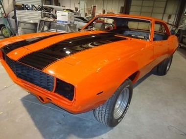 Bright orange classic Camaro with black racing stripes, inside a garage.