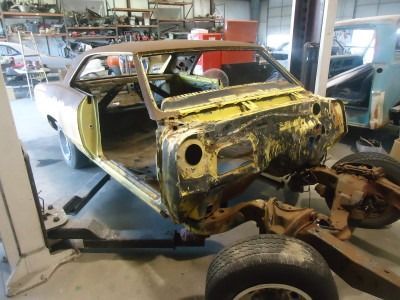 A classic muscle car in a garage undergoing restoration; yellowed body, missing parts, and detached chassis.