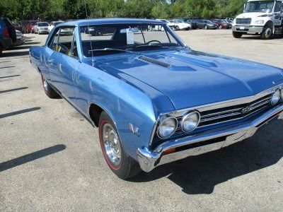 Blue 1967 Chevrolet Chevelle coupe parked on asphalt. It has chrome trim, a hood scoop, and red-line tires.