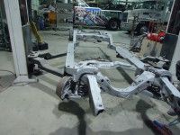 Gray car chassis frame on a shop floor, with suspension components visible.