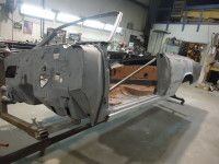 Car frame undergoing restoration in a garage, gray metal, brown interior, on a metal stand.