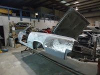 Car body in a repair shop with hood raised. Metal panels show patchwork and primer.